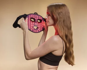 Woman holding BON CHARGE red light therapy mask close to her face, soft red glow illuminating her skin.