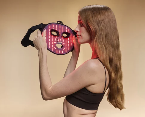 Woman holding BON CHARGE red light therapy mask close to her face, soft red glow illuminating her skin.