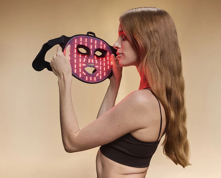 Woman holding BON CHARGE red light therapy mask close to her face, soft red glow illuminating her skin.