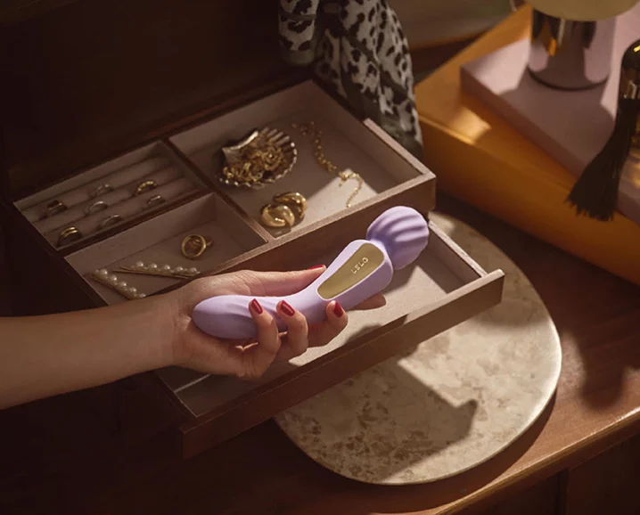 A hand holding the LELO Switch above an open jewelry drawer filled with gold accessories and pearls, styled in a soft, intimate setting.