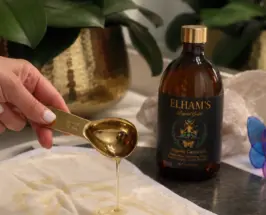 Elham’s Organic Castor Oil in an amber glass bottle beside a gold spoon pouring oil onto a cotton castor oil pack.