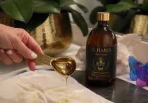 Elham’s Organic Castor Oil in an amber glass bottle beside a gold spoon pouring oil onto a cotton castor oil pack.